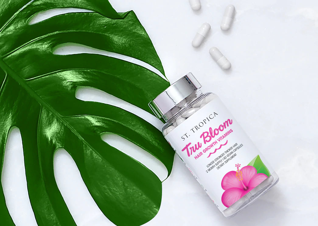 ST. TROPICA hair vitamins on white countertop next to a monstera plant and vitamin capsules