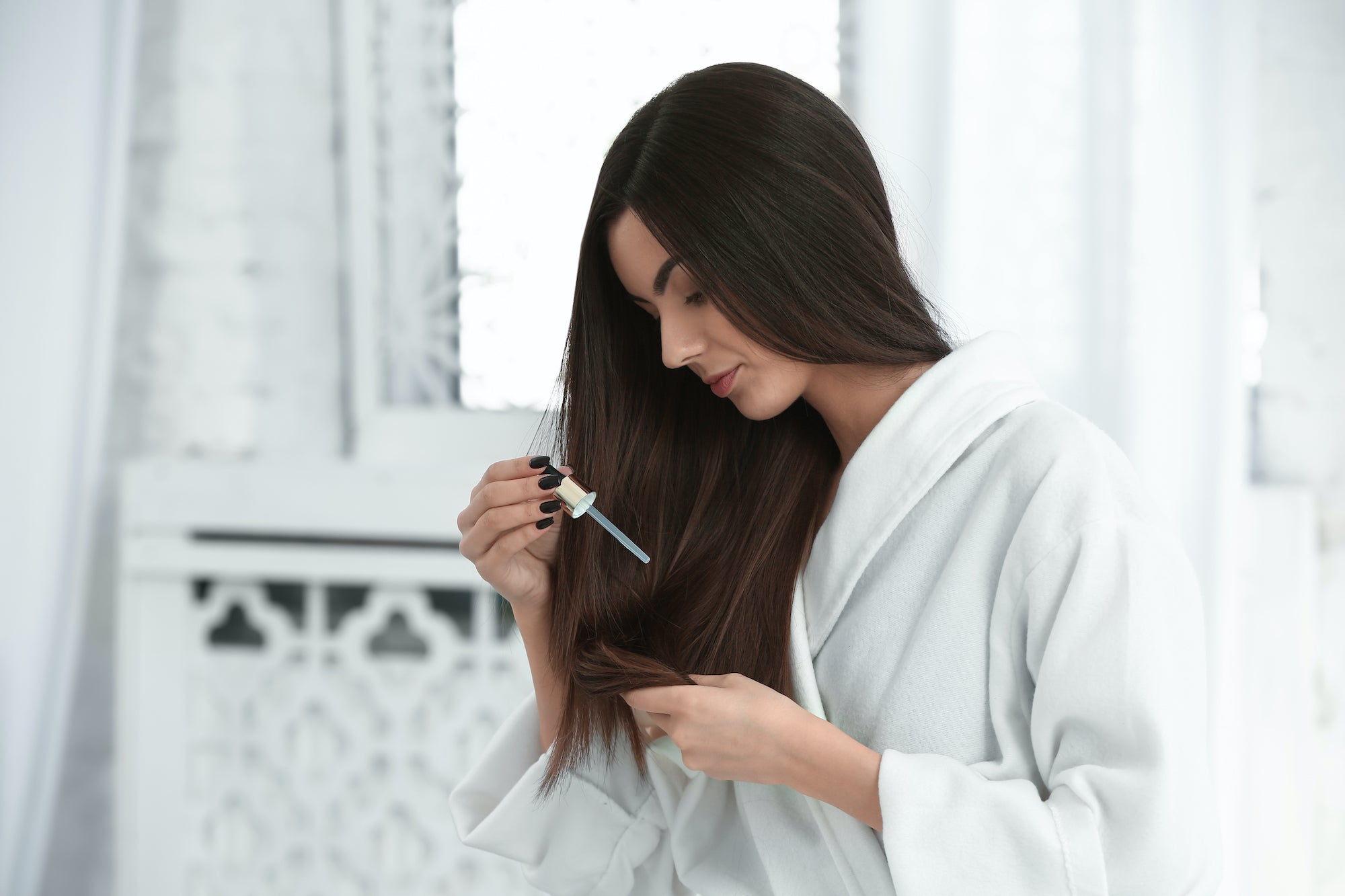 Woman using serum dropper to apply oil to the ends of her dark hair.
