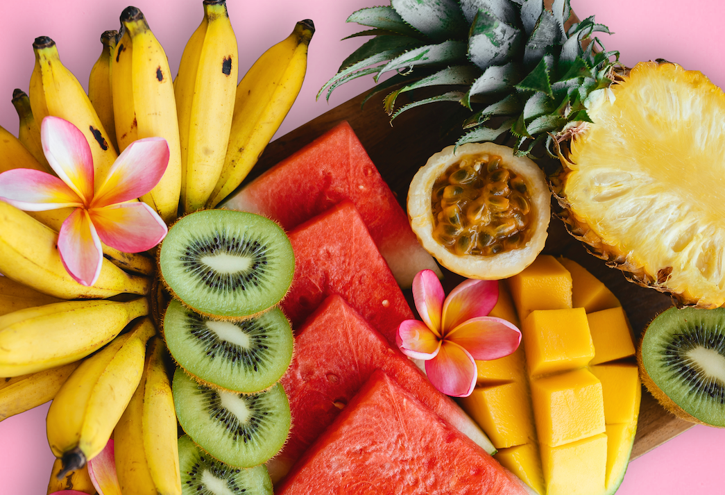 Banana, kiwi, watermelon, mango, kiwi, and pinepple are all laid out on a pink background with tropical flowers placed between some of the fruit.