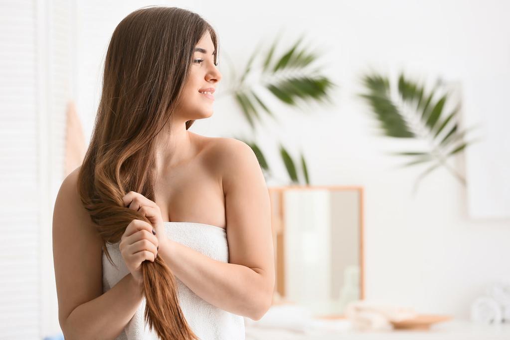 A woman with long brown hair who is standing in her bathroom in a towel is smiling and looking to her side