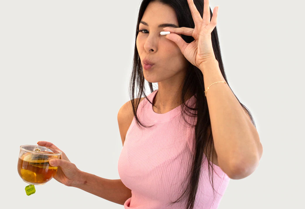 Woman with long black hair holding up vitamin capsule infant of face while puckering mouth. She is holding a cup of green tea in her other hand.