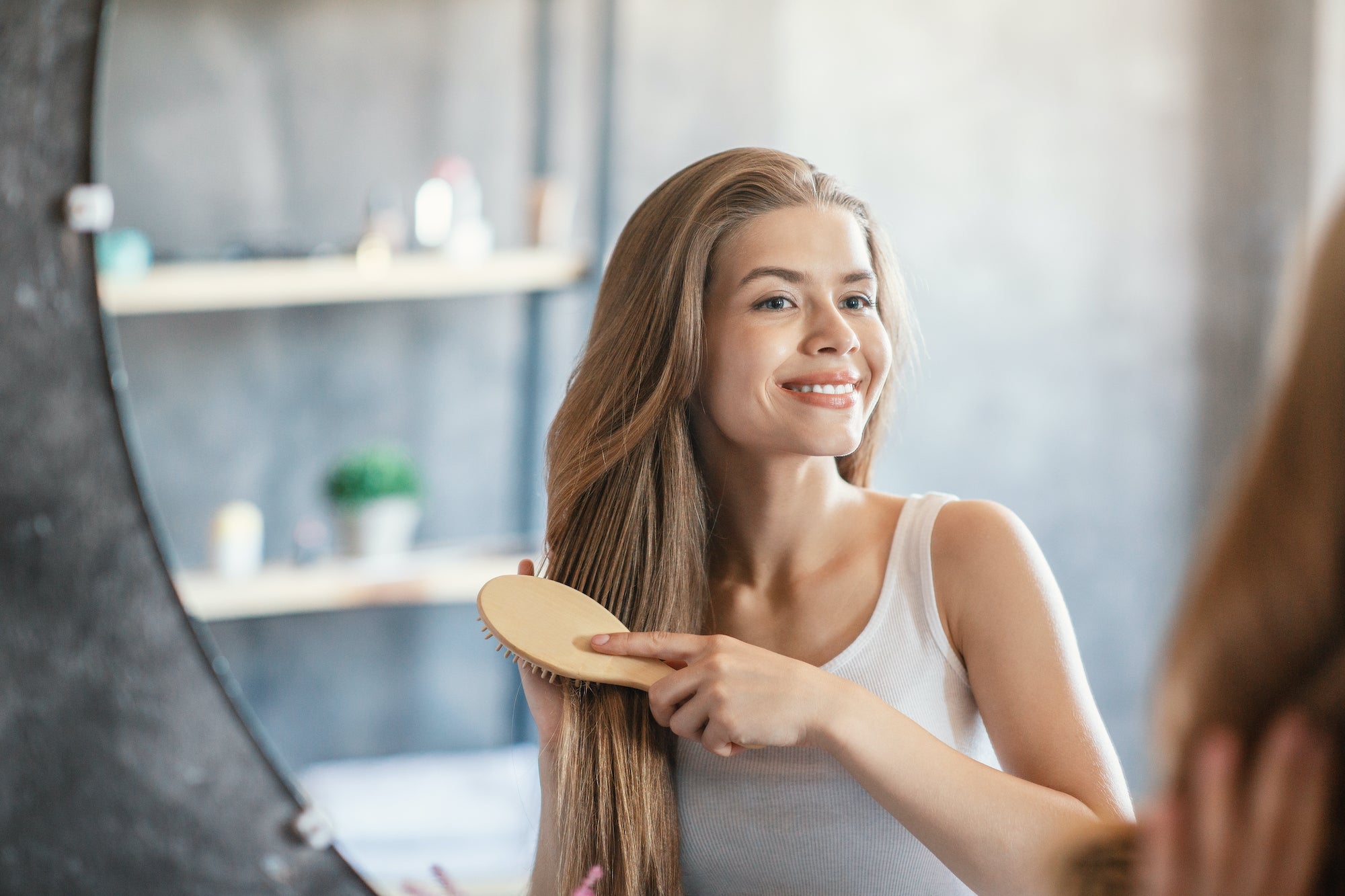 Woman with dark blonde hair looking in mirror and smiling while brushing her hair