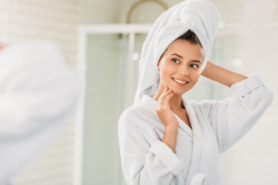 A woman is standing in her bathroom with her hair in a towel and dressed in a robe as she smiles at herself in the mirror. 