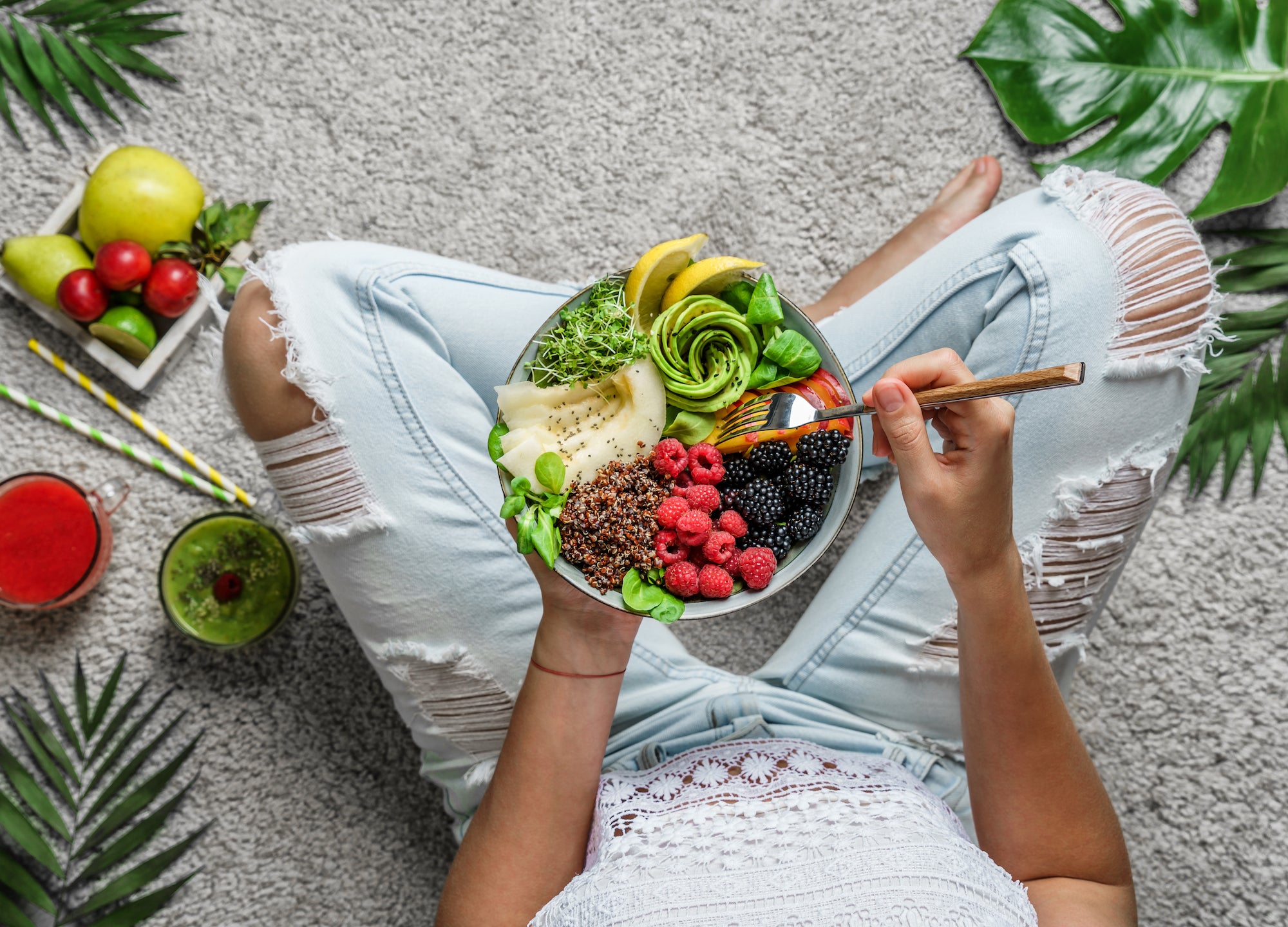A woman who is wearing light jeans and a white top is sitting in the sand and holding a bowl of vibrant fruit and vegetables.
