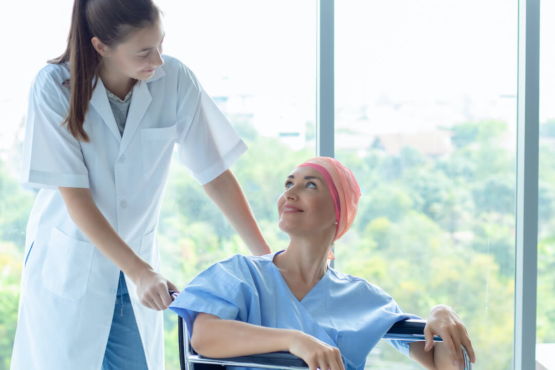 A nurse dressed in scrubs is speaking to a patient in a wheelchair. They are smiling at one another. 