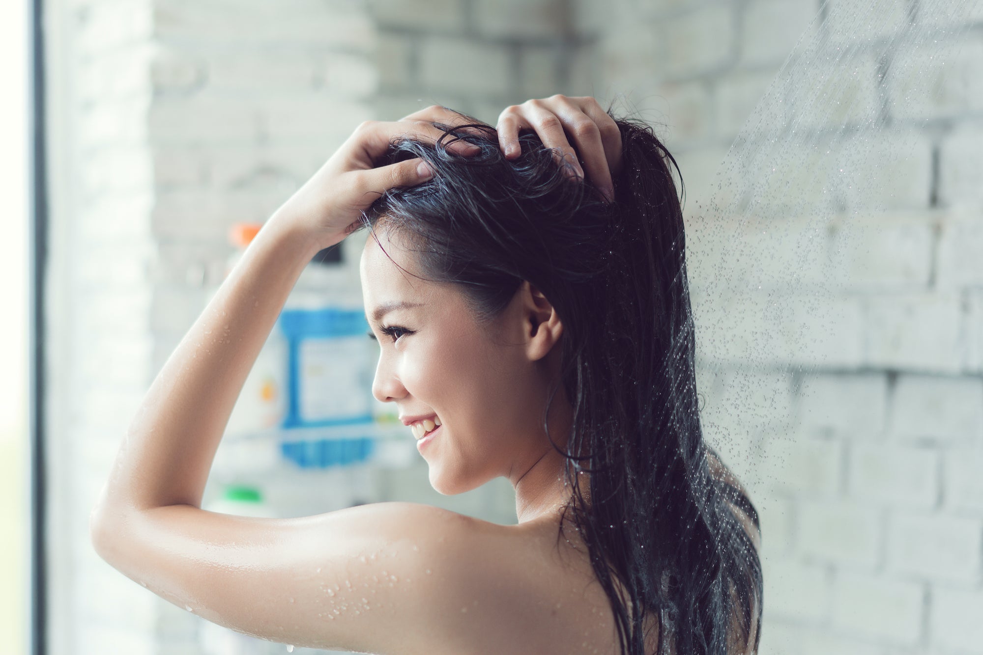Woman with long dark hair smiling and doing a scalp treatment in the shower