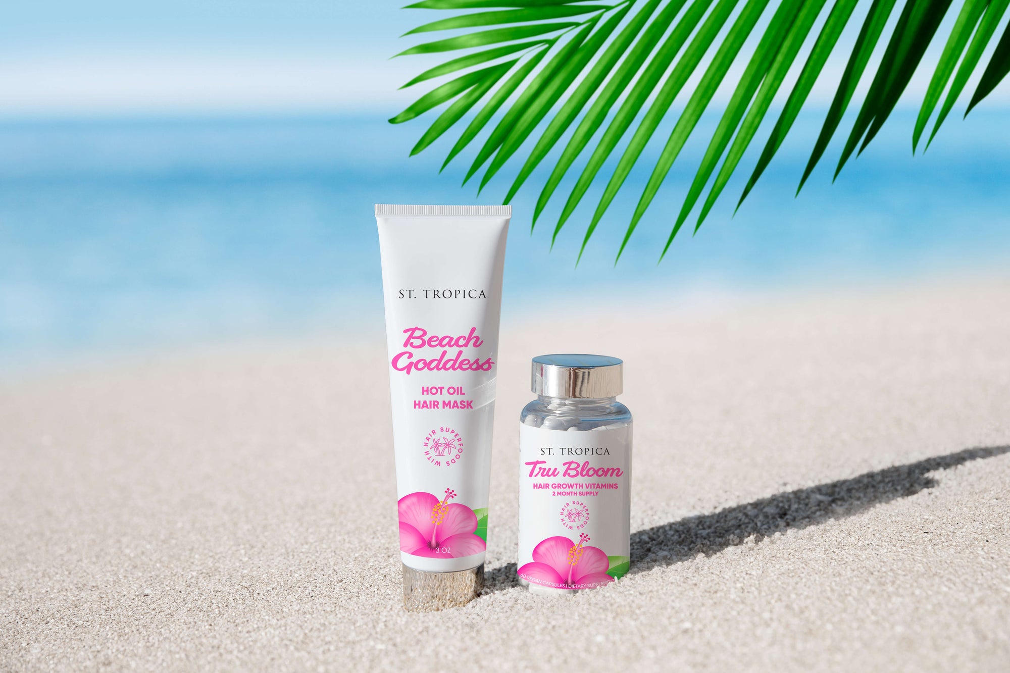ST.TROPICA Hair Vitamins and Beach Goddess Hot Oil Hair Mask flat lay on sandy beach with a pink hibiscus flower in the middle of them.