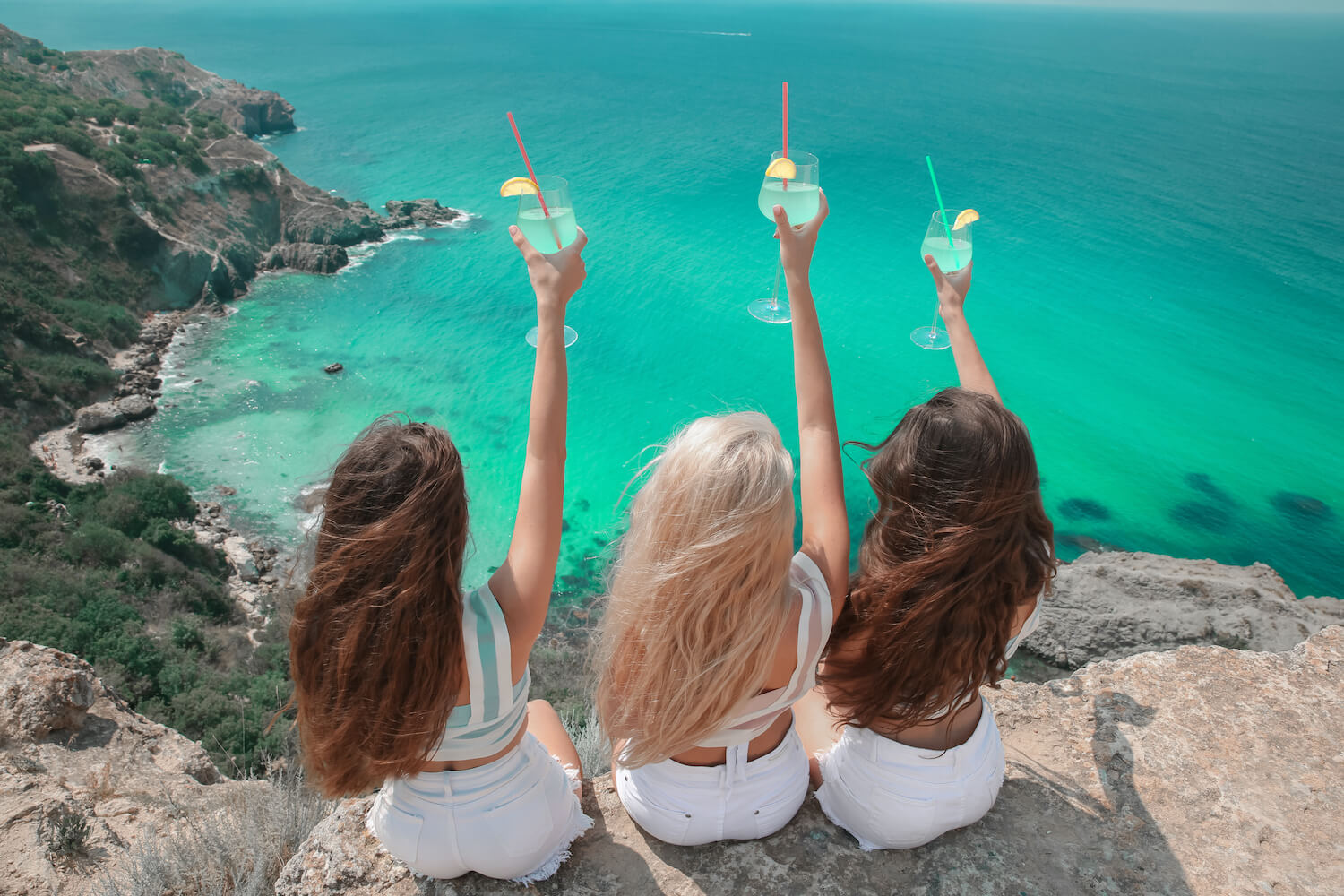 Three women with long healthy hair sitting on a rock overlooking the ocean, holding glasses up to the hair. 